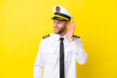 Kafkasyalı uçak pilotu sarı arka planda izole edilmiş bir şekilde elini kulağına koyarak bir şey dinliyor.