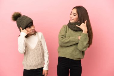 Pembe arka planda izole edilmiş kışlık giysiler giyen küçük kız kardeşler telefon ediyorlar. Bana geri dönüş işareti ver.