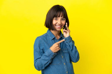 Sarı arka planda izole edilmiş cep telefonu kullanan hamile bir kadın bir ürün sunmak için kenarı işaret ediyor