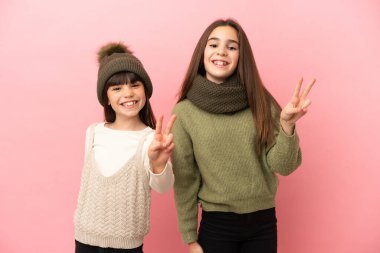 Pembe arka planda izole edilmiş kışlık elbiseler giyen küçük kardeşler gülümsüyor ve zafer işareti gösteriyorlar.