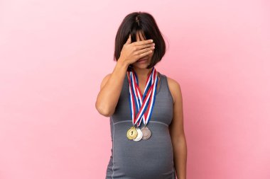 Pregnant mixed race woman with medals isolated on pink background covering eyes by hands. Do not want to see something