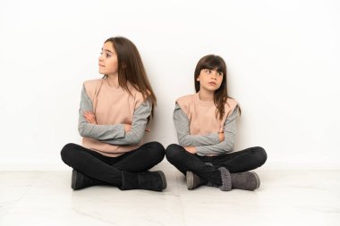 Beyaz arka planda izole edilmiş yerde oturan küçük kız kardeşler dudak ısırırken yüz ifadelerini şaşırtıyorlar.