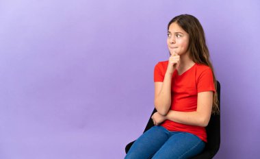 Küçük beyaz kız, mor arka planda izole edilmiş bir sandalyede oturuyor. Şüpheleri var ve düşünüyor.