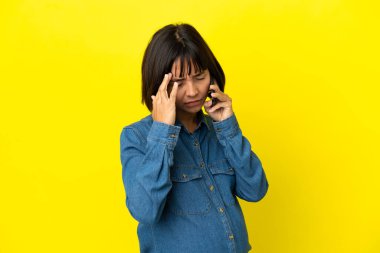 Sarı arka planda izole edilmiş, baş ağrısıyla cep telefonu kullanan hamile bir kadın.