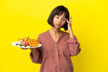 Pregnant woman holding waffles isolated on yellow background with headache