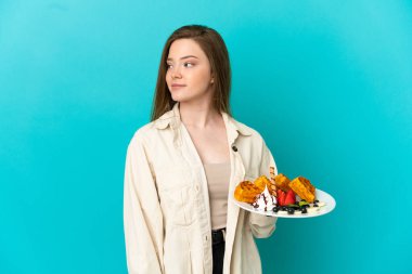 İzole edilmiş mavi arka planda waffle tutan genç kız yan tarafa bakıyor.