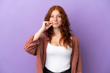 İzole edilmiş mor arka planda genç bir kızıl kız sessizlik işareti gösteriyor.