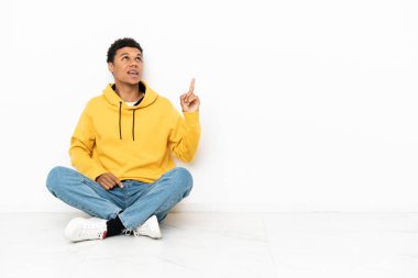 Young African American man sitting on the floor isolated on white background pointing up and surprised