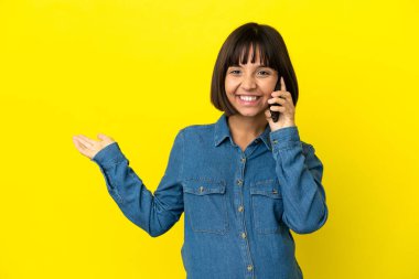 Sarı arka planda izole edilmiş cep telefonu kullanan hamile bir kadın davet edilmek için ellerini yana uzatıyor.