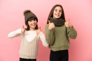 Pembe arka planda izole edilmiş kışlık giysiler giyen küçük kız kardeşler iki eliyle baş parmak işareti yapıyor ve gülümsüyor.