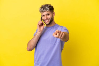 Sarı arka planda izole edilmiş cep telefonu kullanan genç beyaz adam mutlu bir ifadeyle öne bakıyor.
