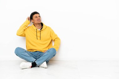 Young African American man sitting on the floor isolated on white background having doubts and with confuse face expression