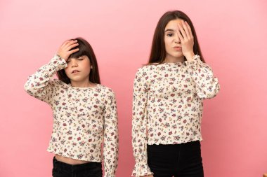 Küçük kız kardeşler pembe arka planda izole edilmiş, şaşırmış ve şok olmuş yüz ifadeleriyle.