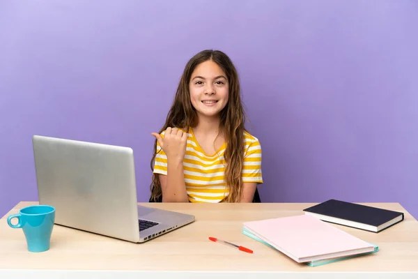 Little student girl in a workplace with a laptop isolated on purple background pointing to the side to present a product