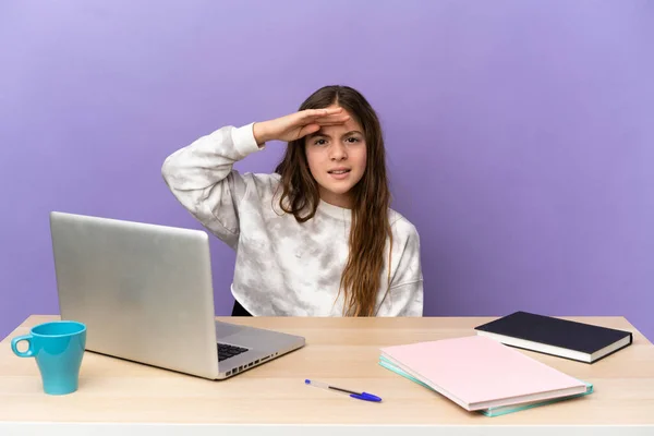 Little student girl in a workplace with a laptop isolated on purple background looking far away with hand to look something