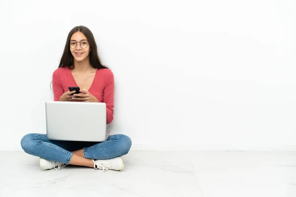 年轻的法国姑娘坐在地板上 带着笔记本电脑 用手机传递信息 — 图库照片
