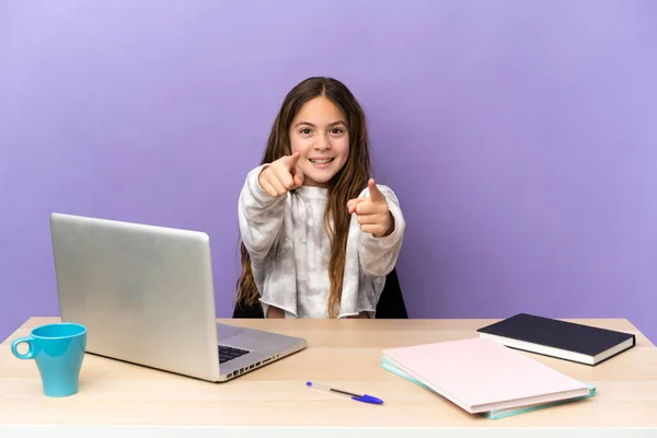 Little student girl in a workplace with a laptop isolated on purple background surprised and pointing front