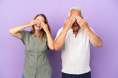 Mor arka planda izole edilmiş orta yaşlı çift gözlerini elleriyle kapatıyor.