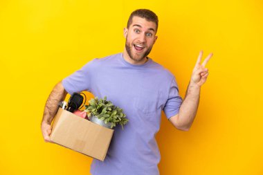 Genç Kafkasyalı, sarı arka planda izole edilmiş şeylerle dolu bir kutuyu alırken gülümsüyor ve zafer işareti gösteriyor.