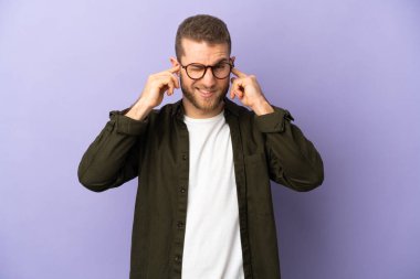 Mor arka planda izole edilmiş yakışıklı beyaz adam hayal kırıklığına uğramış ve kulaklarını kapatmış.