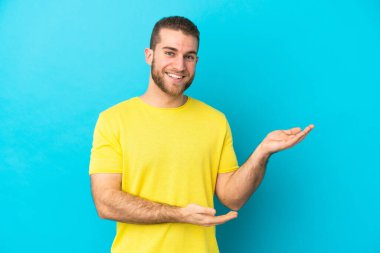 Mavi arka planda izole edilmiş yakışıklı beyaz adam, davet edilmek için ellerini yana uzatıyor.