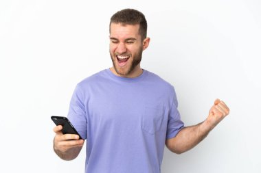 Beyaz arka planda izole edilmiş genç yakışıklı beyaz adam cep telefonu kullanıyor ve zafer jesti yapıyor.
