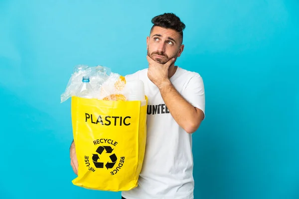 Caucasian man holding a bag full of plastic bottles to recycle isolated on blue background having doubts