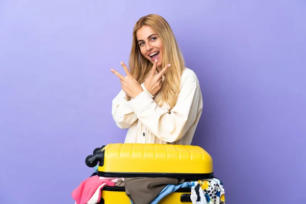 若いですウルグアイブロンド女性とともにスーツケースフルの服上の隔離された紫色の壁笑顔とショー勝利サイン — ストック写真