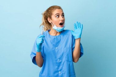 Genç kadın diş hekimi, mavi arka plan üzerinde sürpriz yüz ifadesiyle aletler tutuyor.