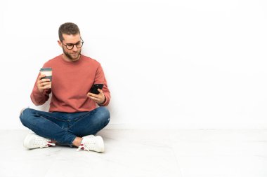 Yerde oturan yakışıklı genç bir adam elinde kahve ve bir cep telefonu tutuyor.