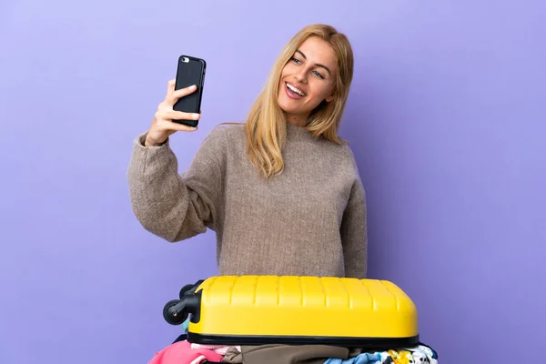 Young Uruguayan blonde woman with a suitcase full of clothes over isolated purple wall making a selfie