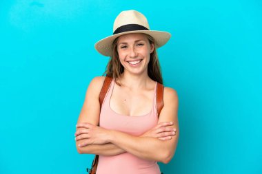 Yaz tatilinde genç beyaz bir kadın mavi arka planda izole edilmiş kollarını ön cephede kavuşturuyor.