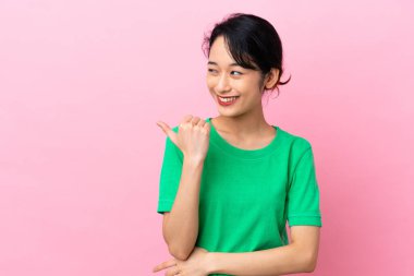 Pembe arka planda izole edilmiş genç Vietnamlı kadın bir ürünü sunmak için kenarı işaret ediyor.