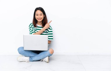 Yerde dizüstü bilgisayarı olan genç Vietnamlı bir kadın beyaz duvarda izole edilmiş bir şekilde oturuyor. Yan tarafı göstererek bir ürün sunuyor.