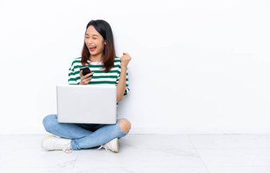 Yerde dizüstü bilgisayarı olan genç Vietnamlı bir kadın zafer pozisyonunda telefonuyla beyaz duvarda izole bir şekilde oturuyor.