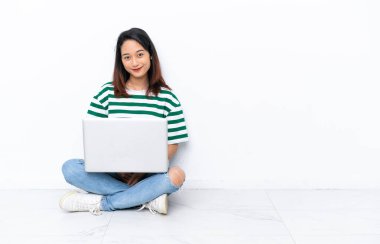 Dizüstü bilgisayarı olan genç Vietnamlı kadın beyaz duvarda izole bir şekilde oturmuş gülüyor.