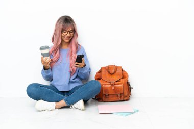 Genç, melez, pembe saçlı bir ırk kadını beyaz arka planda izole bir şekilde oturuyor. Elinde kahve ve cep telefonu var.