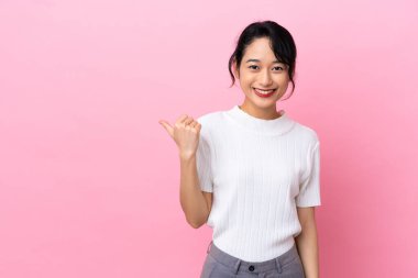 Pembe arka planda izole edilmiş genç Vietnamlı kadın bir ürünü sunmak için kenarı işaret ediyor.
