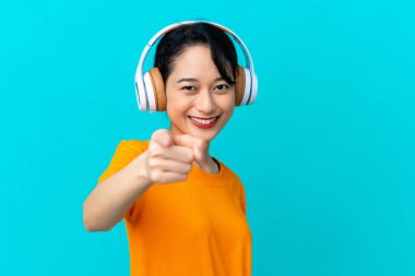 Genç Vietnamlı kadın mavi arka planda izole edilmiş müzik dinliyor ve ön tarafı işaret ediyor.