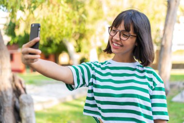Parktaki genç esmer kadın cep telefonuyla selfie çekiyor.