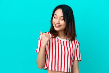 Mavi arka planda izole edilmiş genç Vietnamlı kadın bir ürünü sunmak için kenarı işaret ediyor.