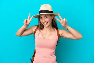 Yaz tatilinde genç beyaz bir kadın mavi arka planda izole bir şekilde zafer işareti gösteriyor.