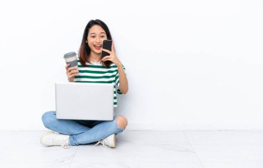 Yerde dizüstü bilgisayarı olan genç Vietnamlı bir kadın beyaz duvarda izole bir şekilde oturuyor. Elinde kahve ve cep telefonu var.