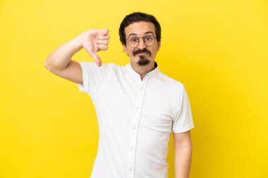 Sarı arka planda izole edilmiş genç beyaz adam olumsuz ifadelerle baş parmağını aşağı gösteriyor.