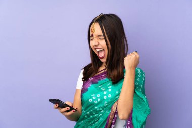 Mor arka planda izole edilmiş genç Hintli kadın telefon zafer pozisyonunda.