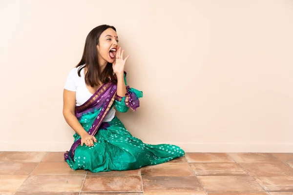 Young Indian woman sitting on the floor shouting with mouth wide open to the side