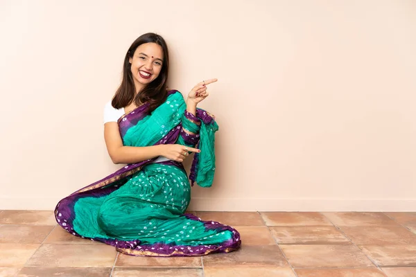 年轻的印度妇女坐在地板上 手指指向旁边 展示着一种产品 — 图库照片