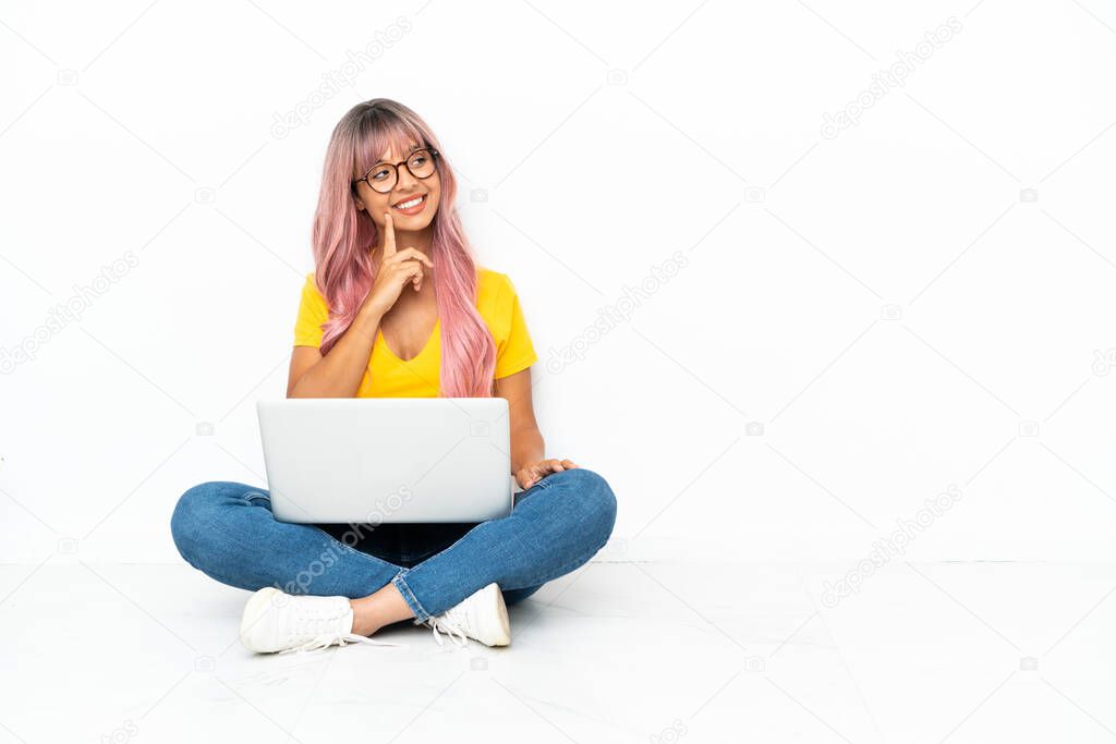 Young mixed race woman with a laptop with pink hair sitting on the floor isolated on white background thinking an idea while looking up