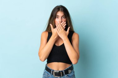 Mavi arka planda izole edilmiş genç beyaz kadın elleriyle ağzını kapatıyor.