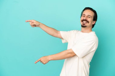 Mavi arka planda izole edilmiş genç beyaz bir adam parmağını yana doğru uzattı ve bir ürün sundu.